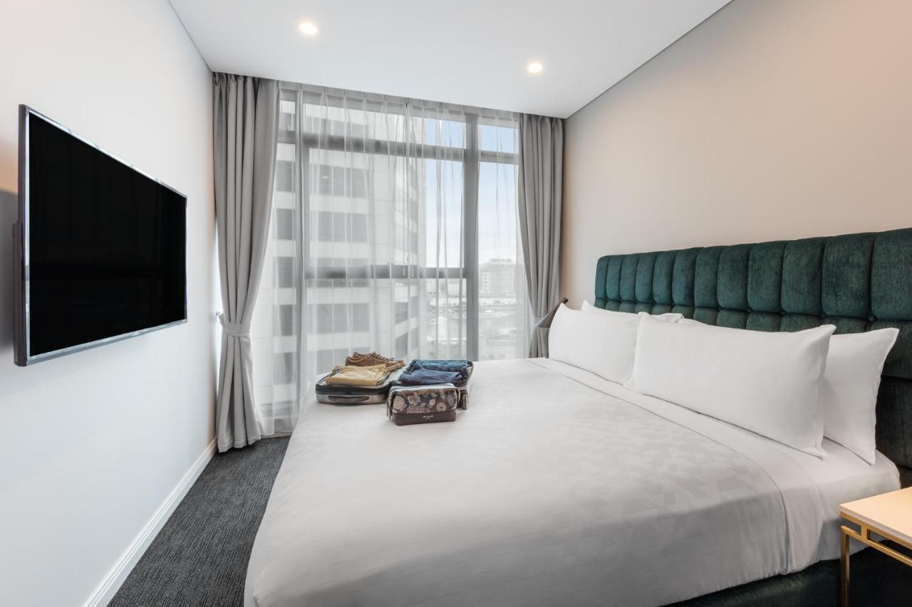 Meriton Suites Sussex Street, Sydney Exterior photo The photo shows a modern hotel bedroom. The room features a large bed with a green upholstered headboard and crisp white linens. There are two pillows on the bed and a few folded items of clothing neatly placed on it. A flat-screen TV is mounted on t