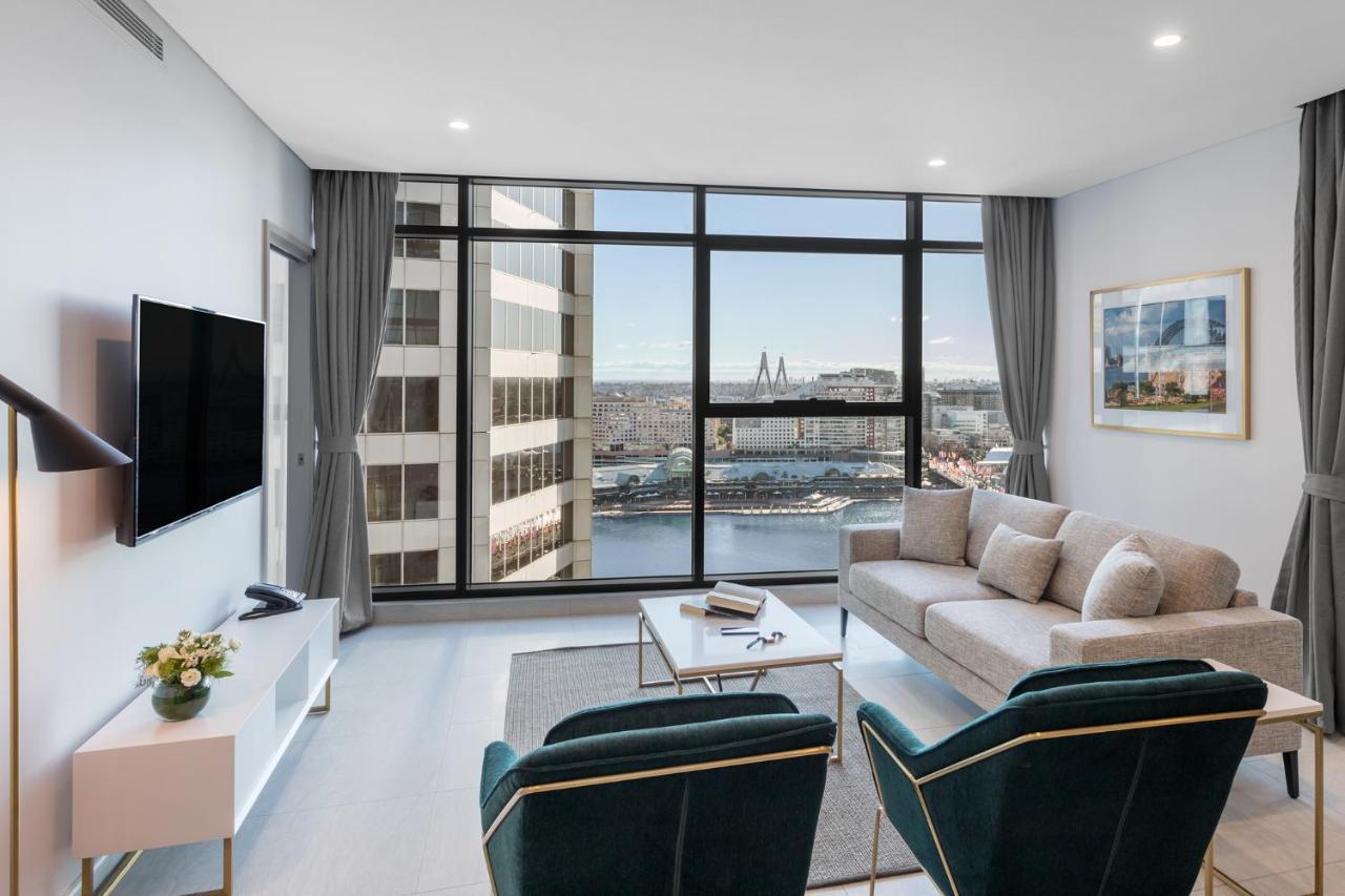 Meriton Suites Sussex Street, Sydney Exterior photo The photo shows a modern living room with large windows that offer a view of the cityscape and water. The interior features a light color palette with pale blue walls and soft lighting. There is a comfortable gray sofa and two green accent chairs, ar