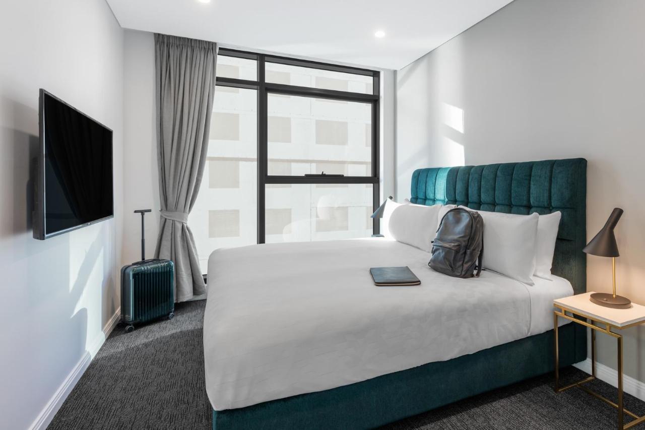 Meriton Suites Sussex Street, Sydney Exterior photo The photo shows a modern hotel room featuring a comfortable bed with white bedding and a teal-colored headboard. There is a neatly placed black backpack on the bed, along with a small notebook or tablet. A television is mounted on the wall opposite t