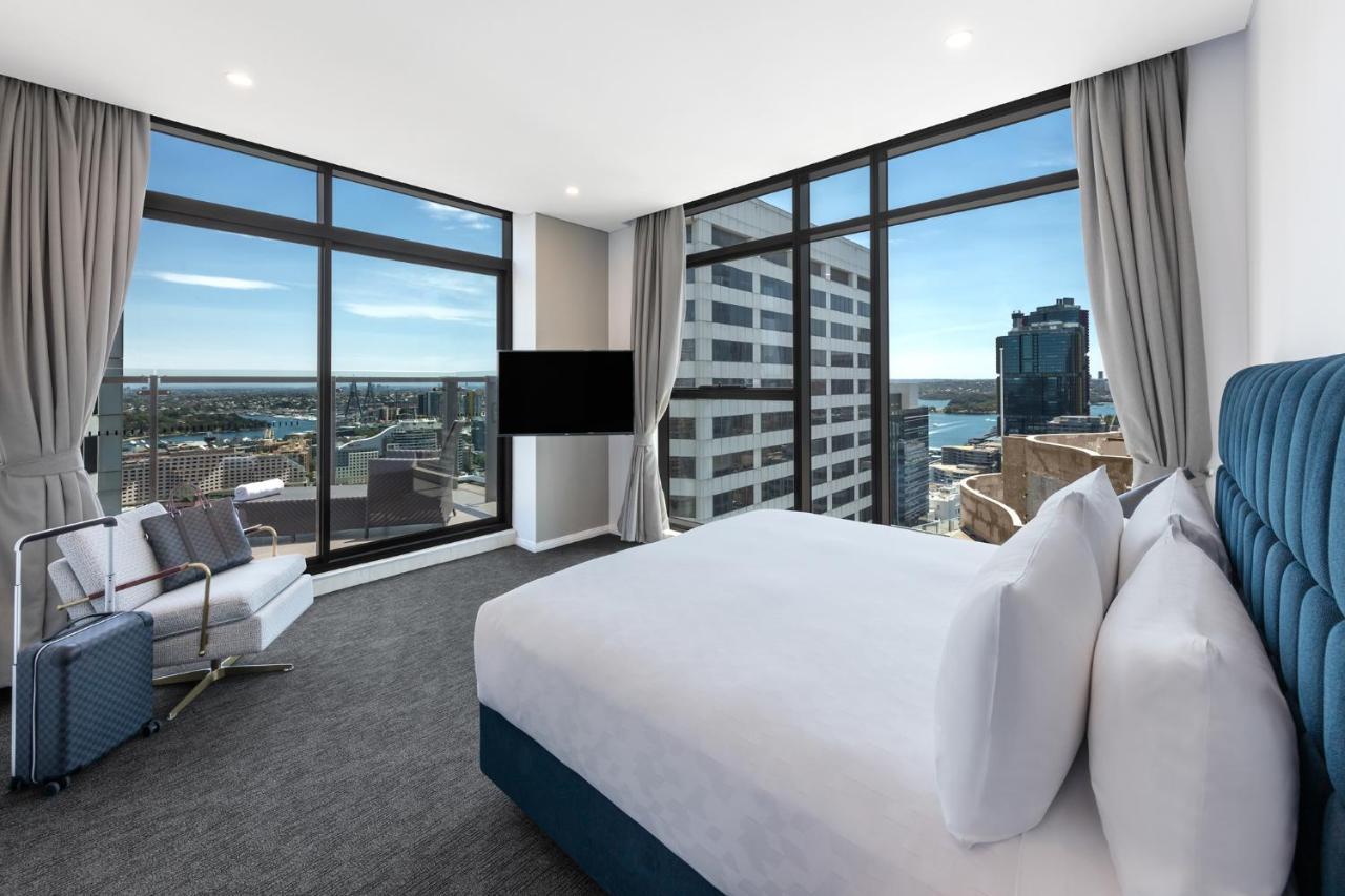 Meriton Suites Sussex Street, Sydney Exterior photo The photo shows a modern hotel room featuring large windows that provide a panoramic view of the cityscape and waterfront. The room is furnished with a king-size bed dressed in white linens, a comfortable armchair, and a flat-screen TV mounted on the