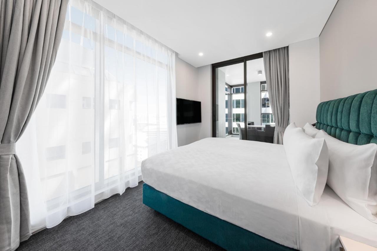 Meriton Suites Sussex Street, Sydney Exterior photo The photo shows a modern bedroom featuring a large bed with a white duvet and pillows. The headboard is turquoise, adding a pop of color to the neutral-toned room. There are sheer white curtains covering large windows that let in natural light, creat