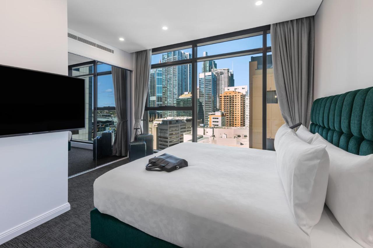 Meriton Suites Sussex Street, Sydney Exterior photo The photo shows a modern hotel room featuring a large bed with white linens and a green upholstered headboard. There is a sleek television mounted on the wall opposite the bed, and the room has large windows that provide a view of a cityscape, includ