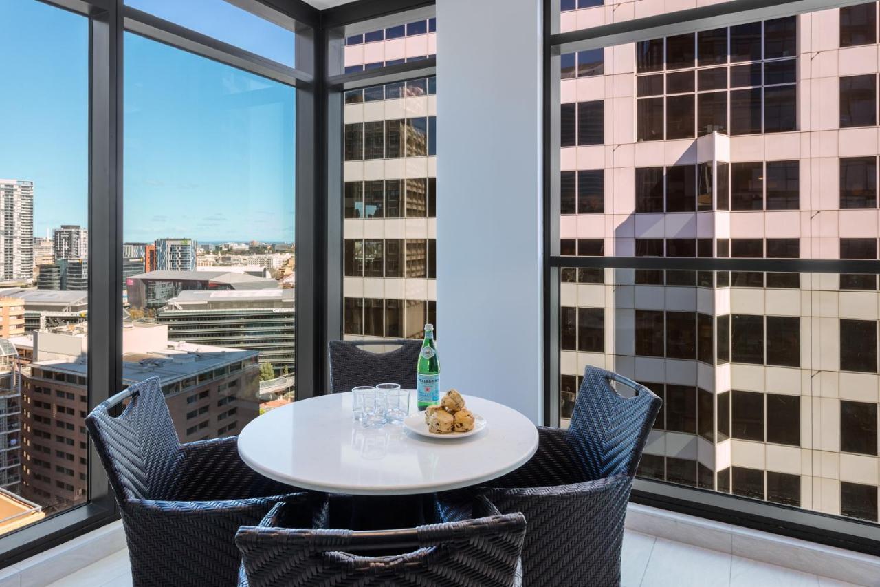 Meriton Suites Sussex Street, Sydney Exterior photo The photo shows a modern dining space with a round white table set for four. There are four black woven chairs around the table. On the table, there's a bottle of sparkling water, two glasses, and a small plate with several cookies. The large windows