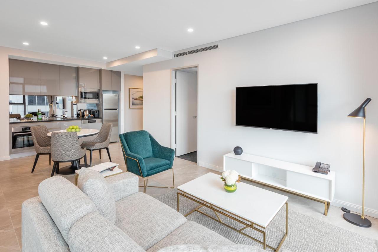 Meriton Suites Sussex Street, Sydney Exterior photo The photo shows a modern living room area. It features a light gray sofa and a coffee table with a decorative plant. There are two chairs, one in teal and a round one, positioned around the coffee table. A flat-screen TV is mounted on the wall, and t