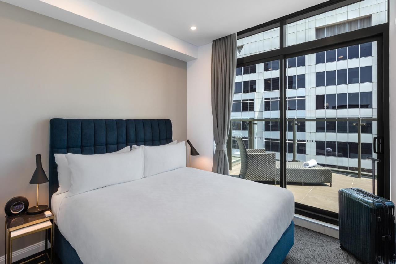 Meriton Suites Sussex Street, Sydney Exterior photo The photo shows a modern hotel room with a large bed featuring a blue upholstered headboard and white linens. Natural light enters through a large window, revealing a view of a building exterior, likely part of the hotel or surrounding area. The room