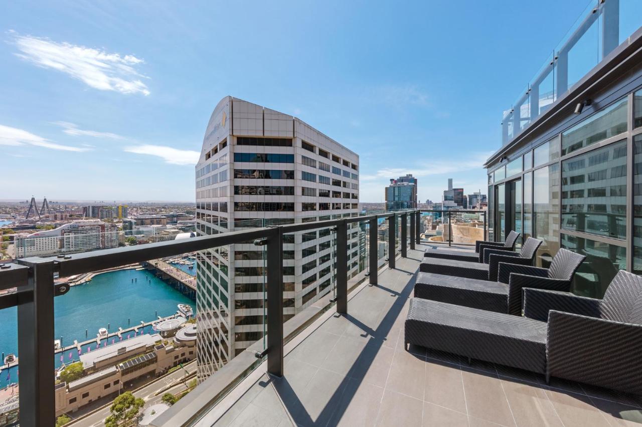 Meriton Suites Sussex Street, Sydney Exterior photo The photo shows a balcony or terrace with a panoramic view of a city skyline. The balcony features modern outdoor furniture, including sleek lounge chairs. In the background, you can see a waterfront area with buildings and a harbor, along with a cle