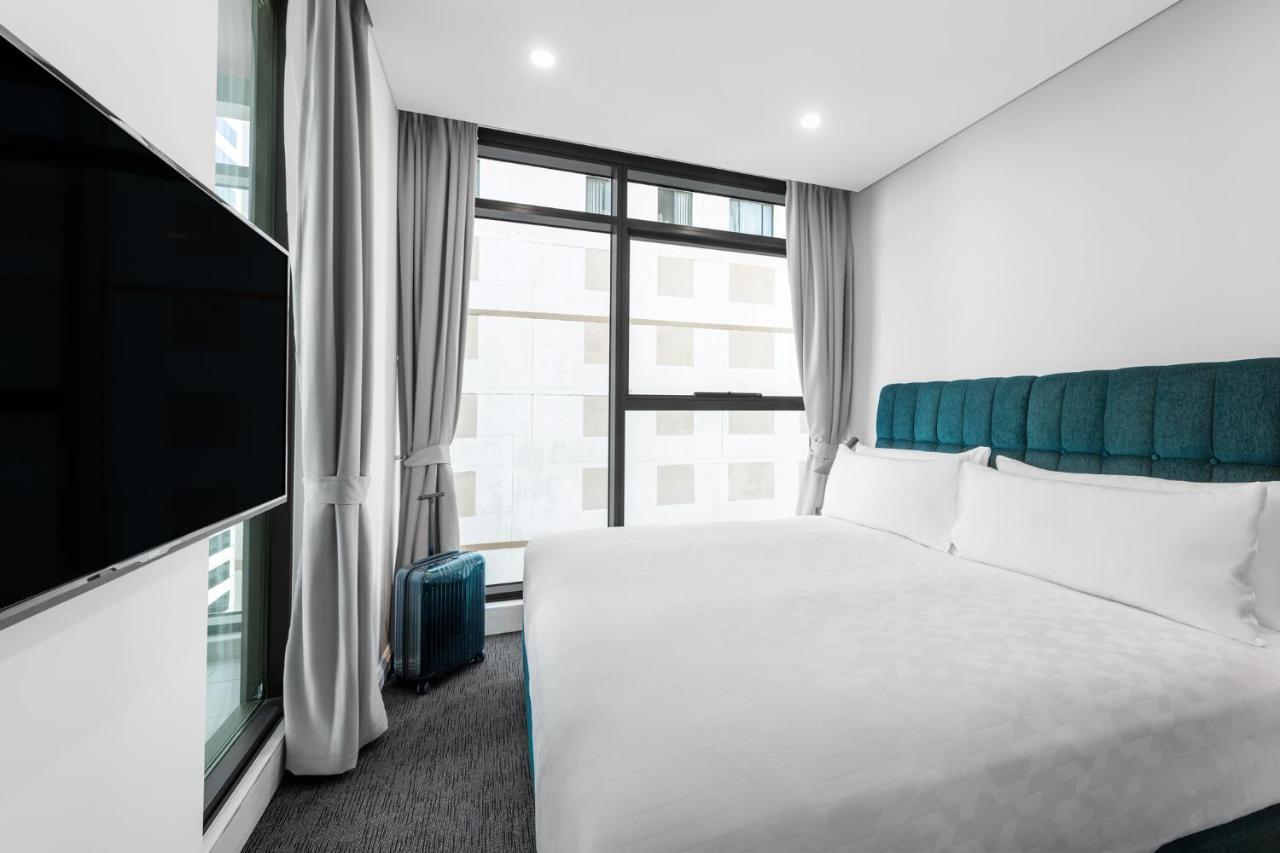 Meriton Suites Sussex Street, Sydney Exterior photo The photo shows a modern hotel room featuring a large bed with a light-colored duvet and a blue upholstered headboard. There are two pillows on the bed. A large window allows natural light to enter the room, and gray curtains frame the window. Next t