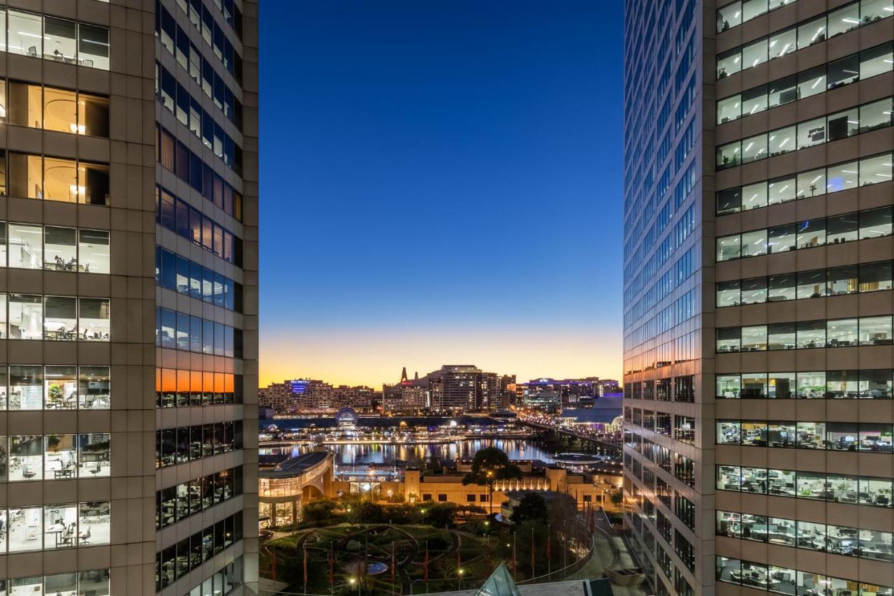 Meriton Suites Sussex Street, Sydney Exterior photo The photo shows a view between two tall buildings, likely office towers, during twilight. On one side, there are glass windows reflecting the evening sky that transitions from blue to orange near the horizon. In the background, a cityscape is visible