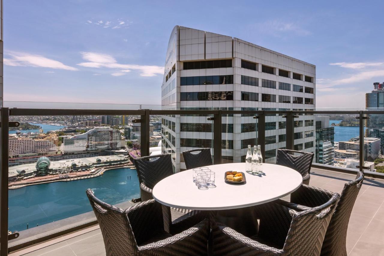 Meriton Suites Sussex Street, Sydney Exterior photo The photo shows a balcony or terrace with a round table and several chairs arranged around it. The table is set with some drinks and a plate, possibly containing food. In the background, there is a view of a waterway and several buildings, including 