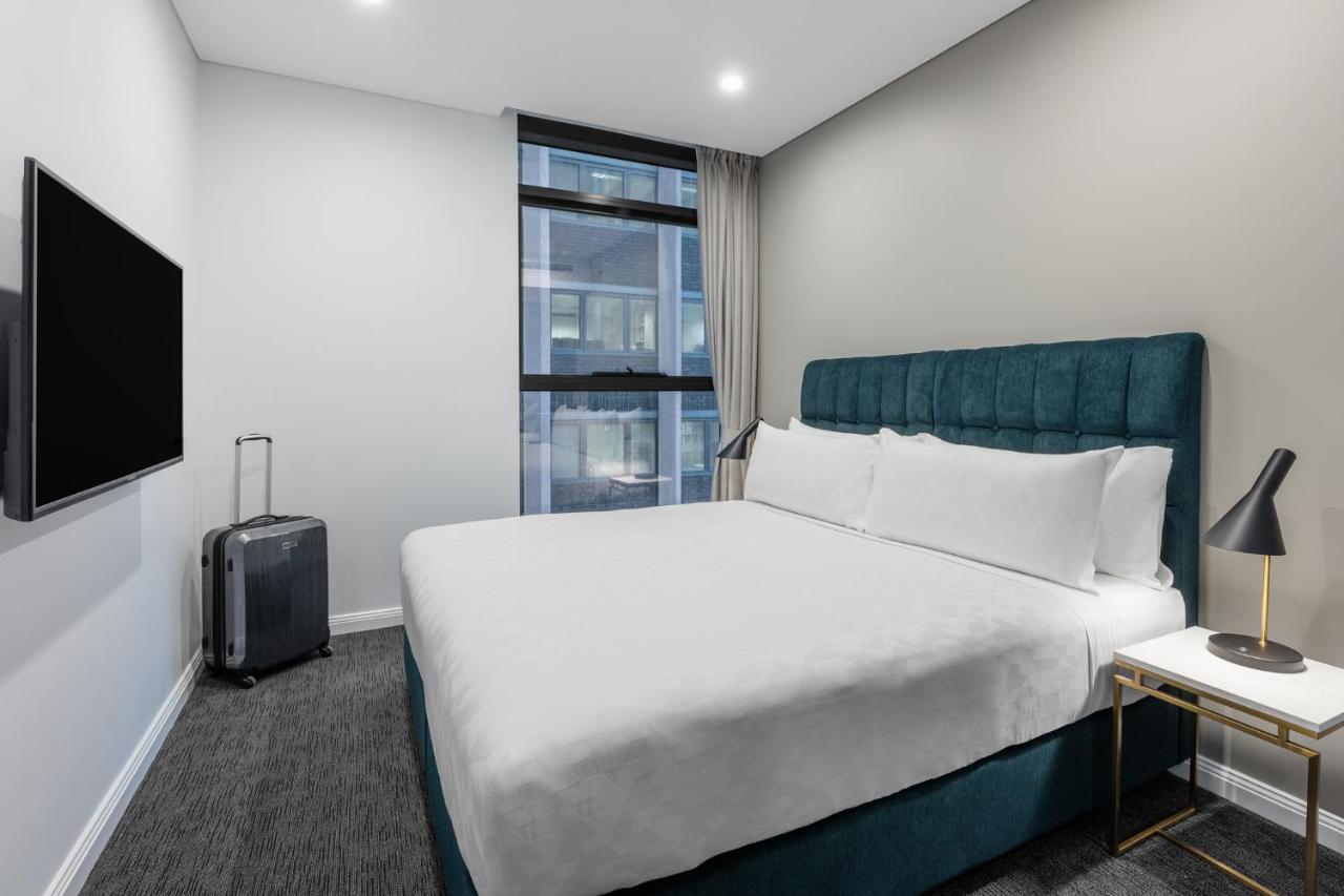 Meriton Suites Sussex Street, Sydney Exterior photo The photo shows a modern hotel room with a clean and minimalist design. There is a large, neatly made bed with white bedding and a teal-colored headboard. On one side of the bed, there is a small nightstand with a lamp. A sleek television is mounted 