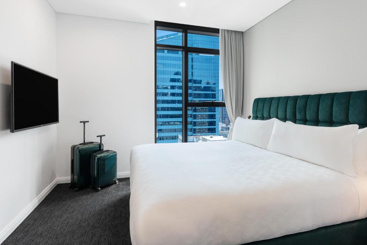 Meriton Suites Sussex Street, Sydney Exterior photo The photo shows a modern hotel room featuring a large bed with white bedding and a green upholstered headboard. There are two green suitcases placed beside the bed. A flat-screen TV is mounted on the wall opposite the bed. The room has a large window