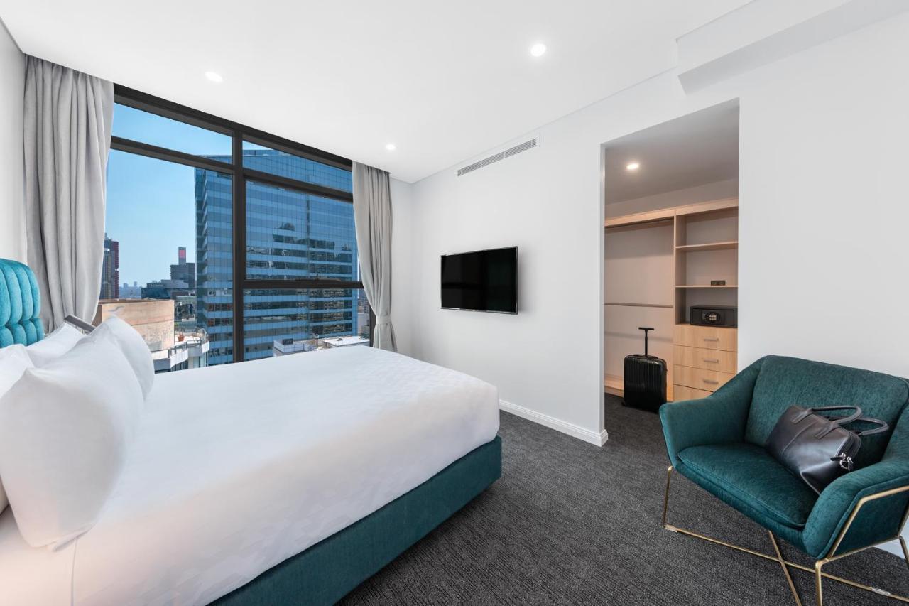 Meriton Suites Sussex Street, Sydney Exterior photo The photo shows a modern hotel room. It features a large window that allows natural light to fill the space, offering a view of the city skyline. The room includes a neatly made bed with a white duvet and a teal-colored base. There is a small green a