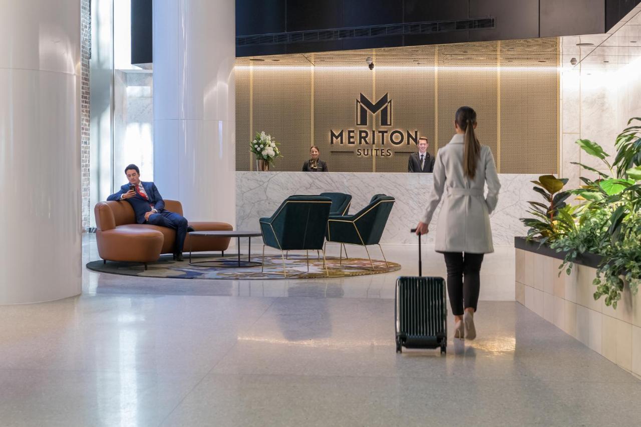 Meriton Suites Sussex Street, Sydney Exterior photo The photo depicts a modern hotel lobby. In the foreground, a woman is walking towards the reception desk, pulling a suitcase behind her. She is dressed in a light-colored coat. To her left, there’s a comfortable seating area with a person sitting on 