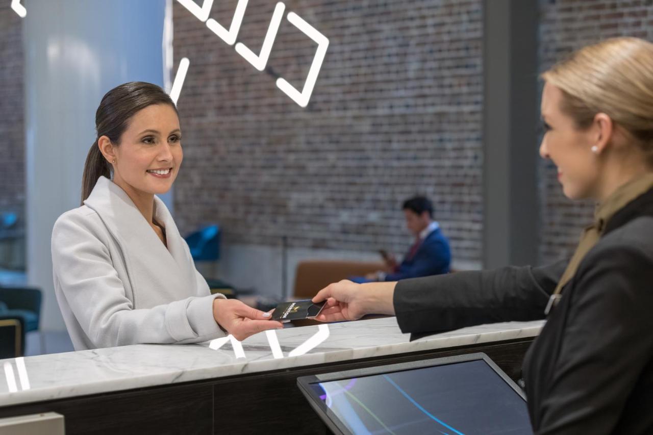 Meriton Suites Sussex Street, Sydney Exterior photo The image depicts a scene at a hotel reception desk. A woman with long hair, dressed in a white outfit, is smiling as she hands her card to a receptionist, who is in a black uniform. The reception area features a modern design, and there is a person 