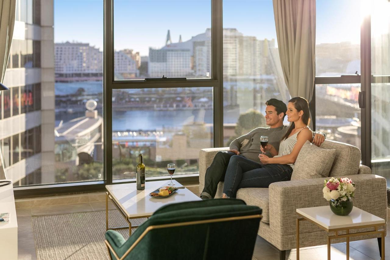 Meriton Suites Sussex Street, Sydney Exterior photo The photo shows a cozy indoor setting with a couple sitting closely together on a light-colored sofa. They are holding drinks, likely wine, and appear to be enjoying each other's company while looking out of a large window. The window provides a view