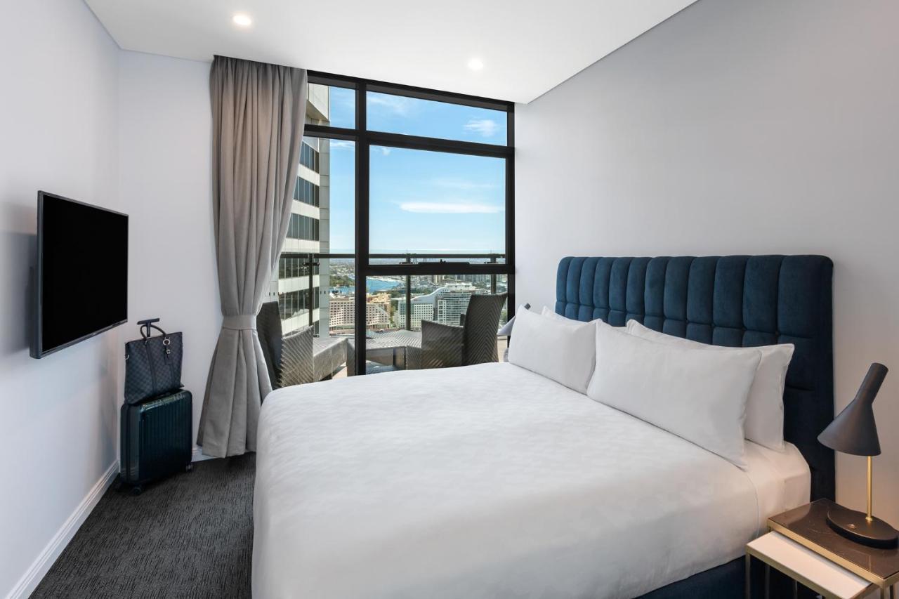 Meriton Suites Sussex Street, Sydney Exterior photo The photo shows a modern hotel room featuring a double bed with white linens and a tufted blue headboard. There is a small bedside table next to the bed. A large window allows natural light to fill the room, offering a view of the city outside, inclu