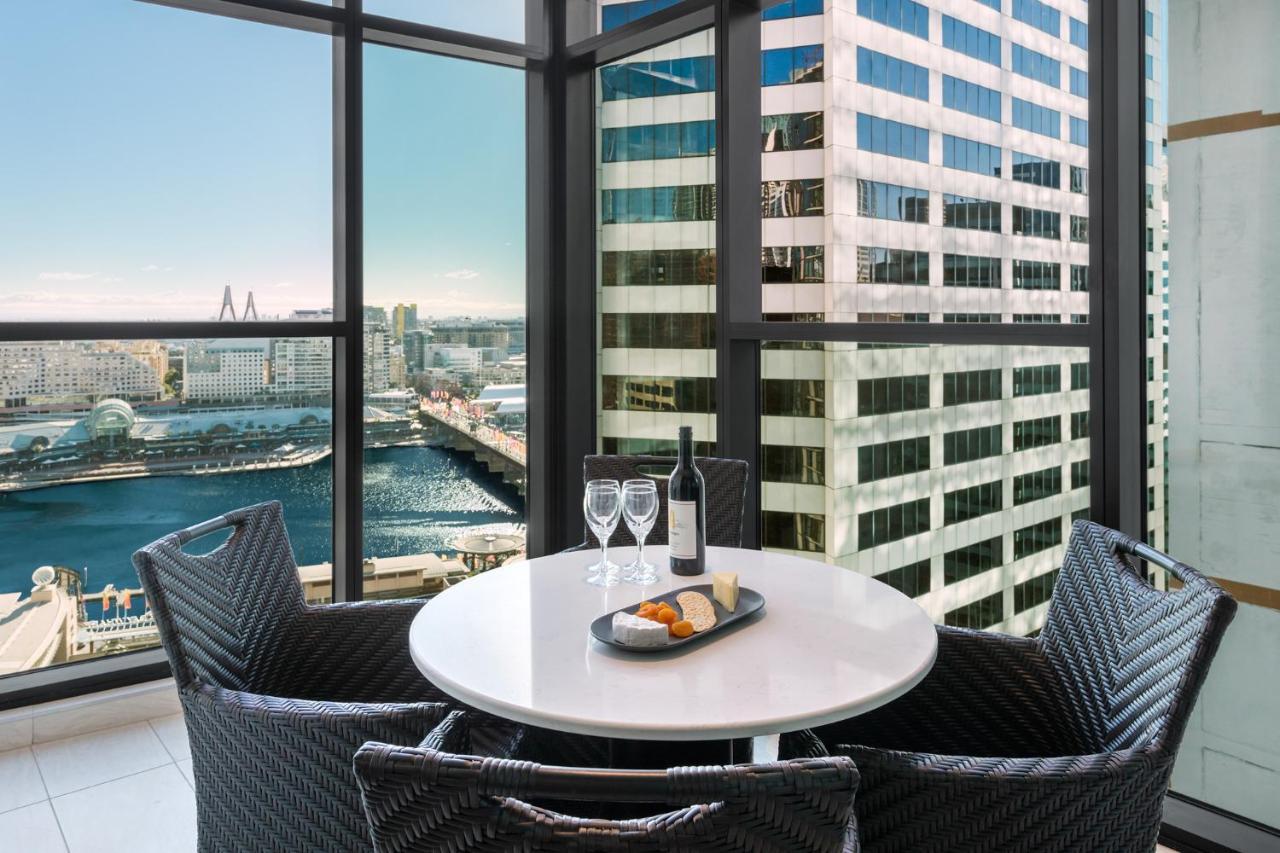 Meriton Suites Sussex Street, Sydney Exterior photo The photo shows a modern dining area with a round table and four woven chairs. On the table, there are two wine glasses, a bottle of wine, and a plate with food items. The large windows provide a view of a cityscape featuring tall buildings and a bod