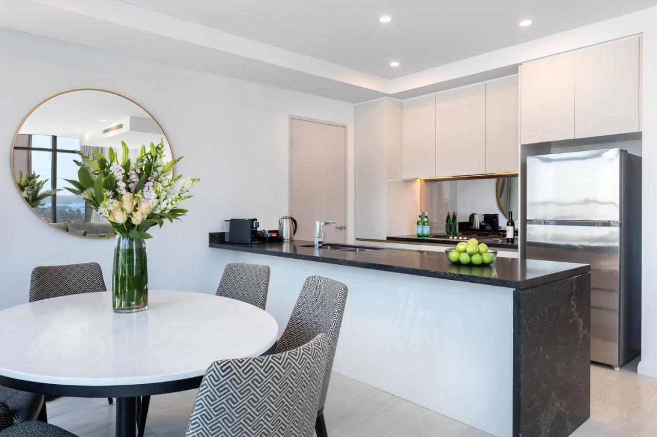 Meriton Suites Sussex Street, Sydney Exterior photo The photo shows a modern kitchen and dining area. In the foreground, there's a circular dining table surrounded by four stylish chairs. A large vase filled with fresh flowers is placed on the table, adding a touch of color and elegance. 

In the kitc