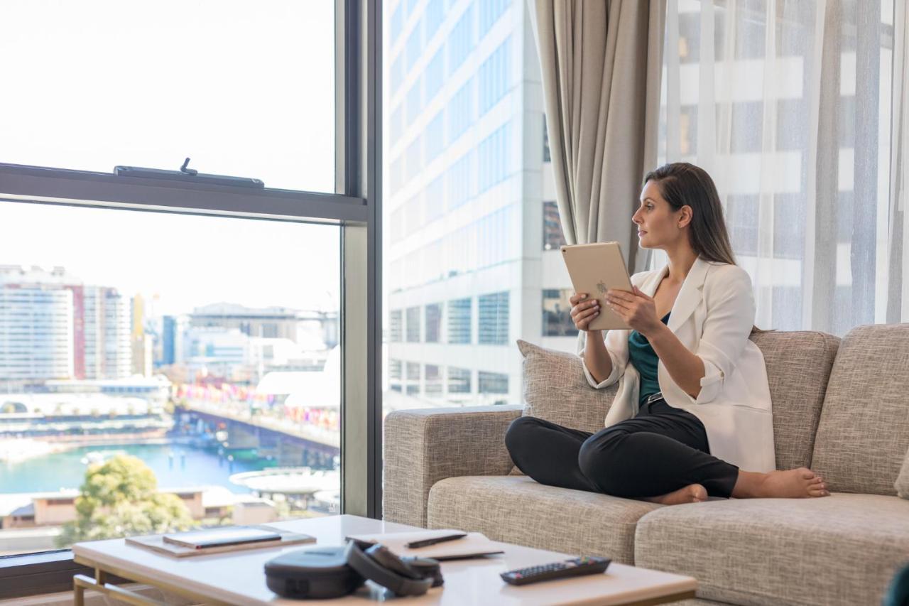 Meriton Suites Sussex Street, Sydney Exterior photo The image portrays a woman sitting cross-legged on a light-colored sofa, engaged with a tablet in her hands. She is dressed in a white blazer and dark pants, appearing relaxed and focused. The setting features large windows that provide a view of a w