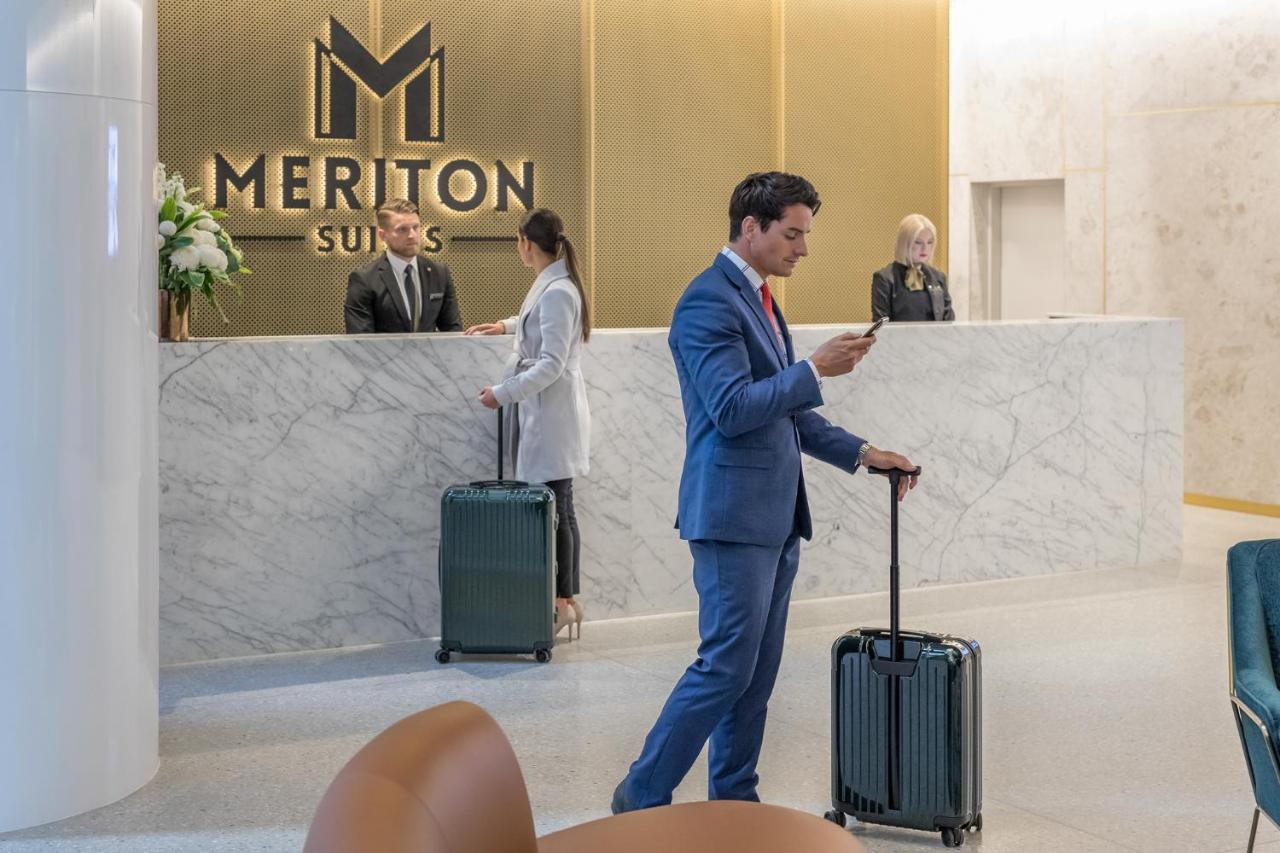 Meriton Suites Sussex Street, Sydney Exterior photo The photo shows the interior of a hotel reception area, specifically at Meriton Suites. In the foreground, a man in a blue suit is standing with a suitcase, looking at his phone. Behind him, there are two staff members at the reception desk, one of w