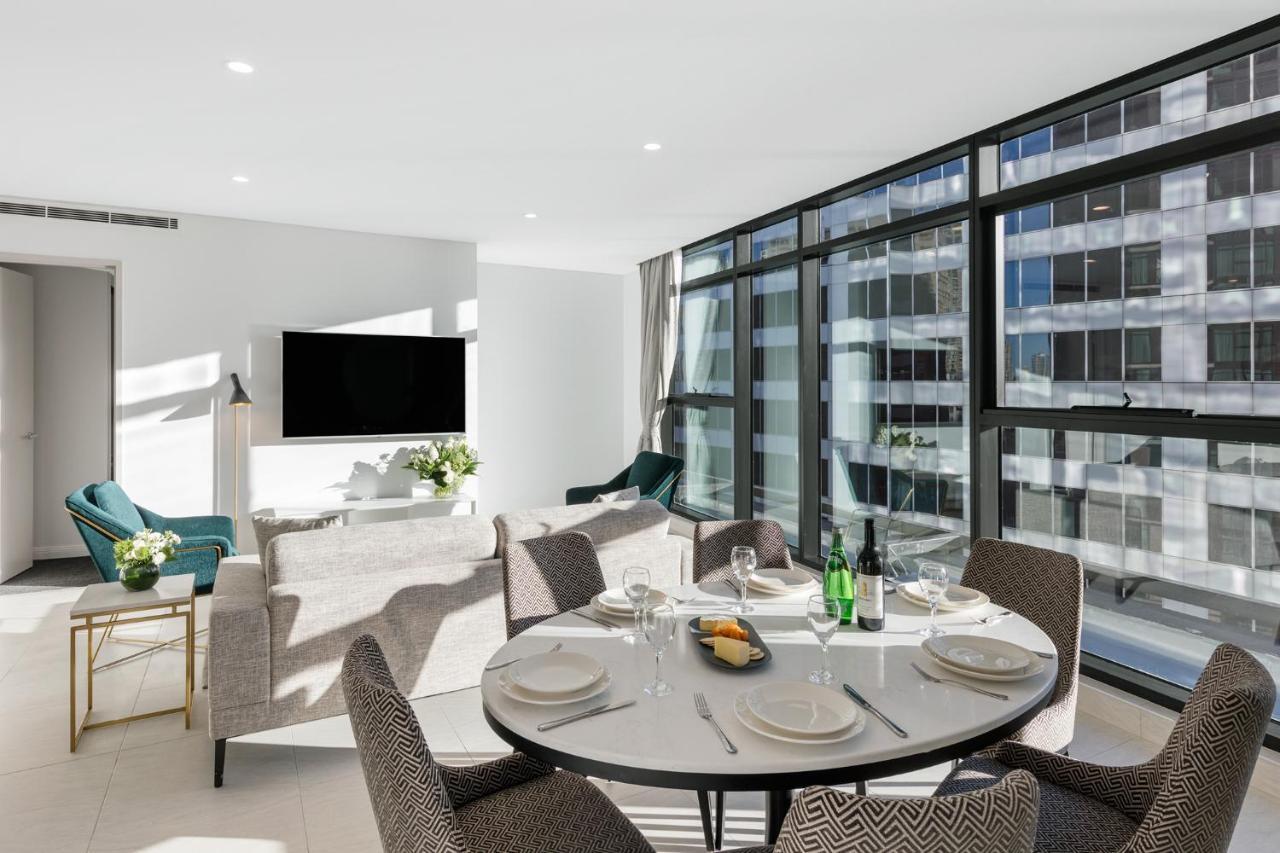 Meriton Suites Sussex Street, Sydney Exterior photo The photo shows a modern and stylish living room and dining area. In the foreground, there is a round dining table set for a meal with plates, cutlery, and drinks. The seating arrangement includes comfortable chairs around the table. 

In the backgro