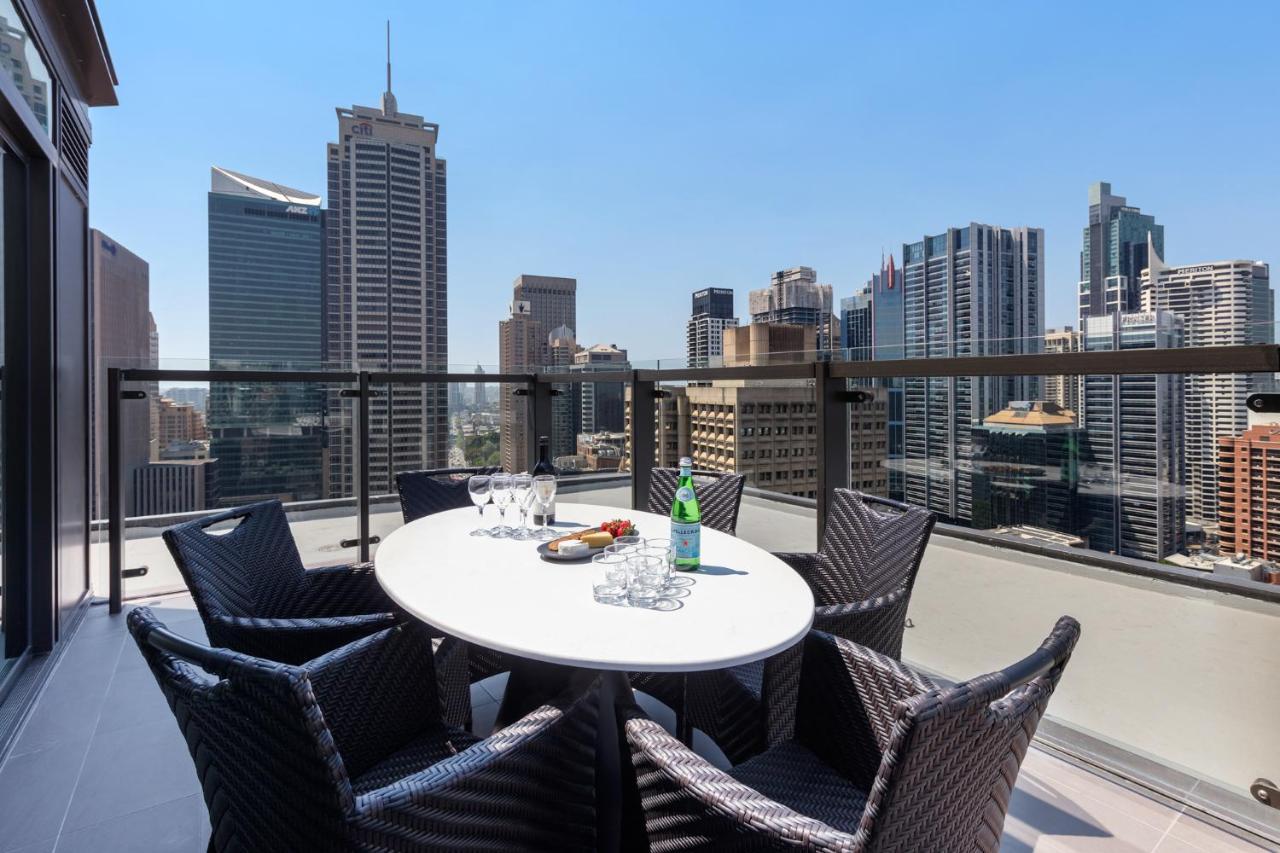 Meriton Suites Sussex Street, Sydney Exterior photo The photo shows a stylish outdoor dining area on a balcony with a round white table surrounded by black wicker chairs. On the table, there are several glasses, a bottle of sparkling water, and a plate of snacks. In the background, there is a panorami