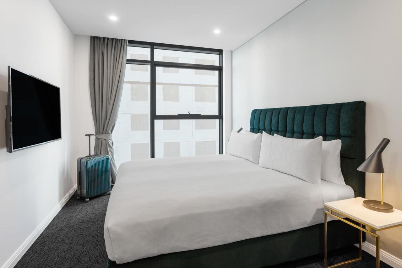 Meriton Suites Sussex Street, Sydney Exterior photo The photo shows a modern hotel room. It features a large bed with a dark green tufted headboard and multiple white pillows. There's a nightstand with a lamp beside the bed. A sleek television is mounted on the wall opposite the bed, and a suitcase is