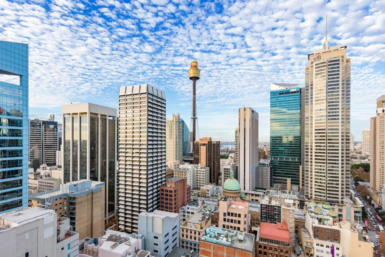 Meriton Suites Sussex Street, Sydney Exterior photo The photo shows a panoramic view of a city skyline dominated by tall skyscrapers. In the center, there is a notable tower with a distinctive golden structure at its top, which is likely a landmark of the city. The buildings have a mix of modern glass