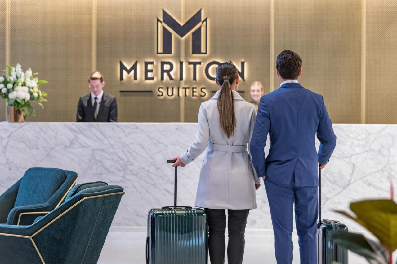 Meriton Suites Sussex Street, Sydney Exterior photo The photo shows a couple standing at a reception area in a hotel named "Meriton Suites." They are both facing the reception desk, where staff members are visible. The man is dressed in a suit, and the woman is wearing a light-colored coat. They each 