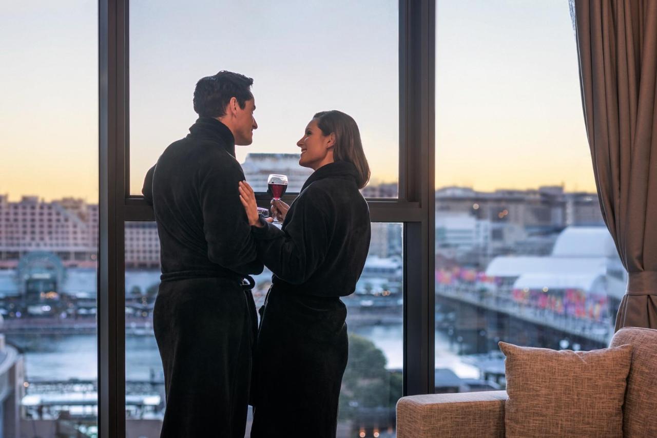 Meriton Suites Sussex Street, Sydney Exterior photo The photo depicts a couple standing closely together by a large window. They are wearing dark bathrobes and holding drinks, gazing into each other’s eyes. The background features a scenic view, likely of a cityscape at sunset, with soft light illumin