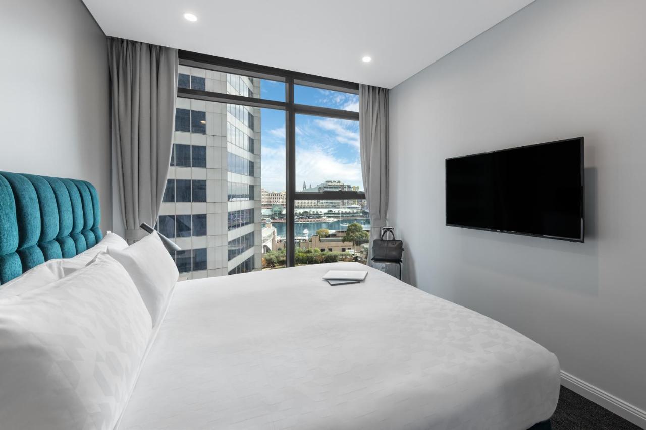 Meriton Suites Sussex Street, Sydney Exterior photo The photo shows a modern hotel room with a large bed featuring a white comforter and pillows. There is a blue headboard that adds a pop of color to the decor. A large window provides a view of the outside, which includes buildings and a glimpse of wa