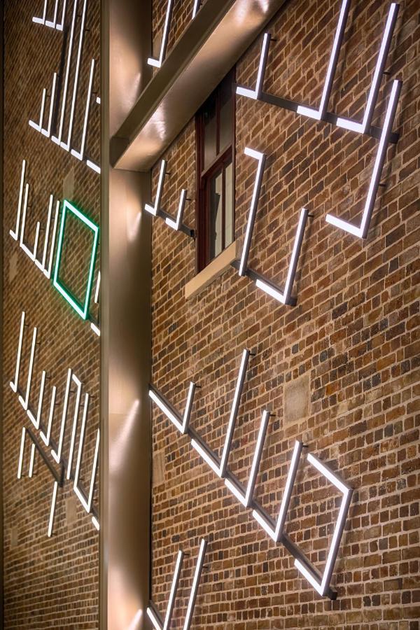 Meriton Suites Sussex Street, Sydney Exterior photo The photo shows a brick wall featuring a design made up of linear light fixtures. These light strips create geometric shapes and patterns, with some sections illuminated in white and one section highlighted in green. The combination of the brick text