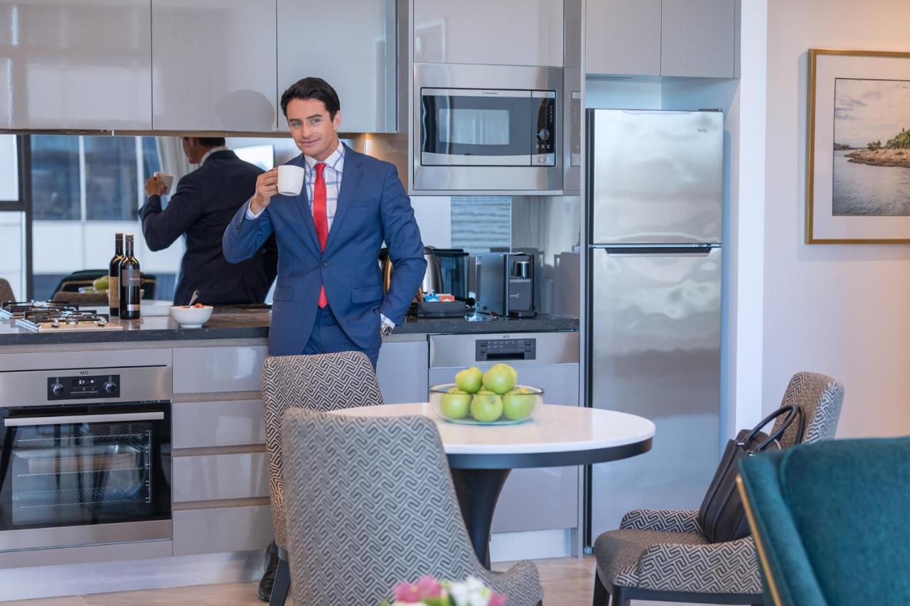 Meriton Suites Sussex Street, Sydney Exterior photo The photo depicts a well-dressed man in a suit standing in a modern kitchen. He is holding a cup, possibly of coffee, and appears to be looking towards the camera. The kitchen features contemporary appliances, including a microwave and refrigerator. 