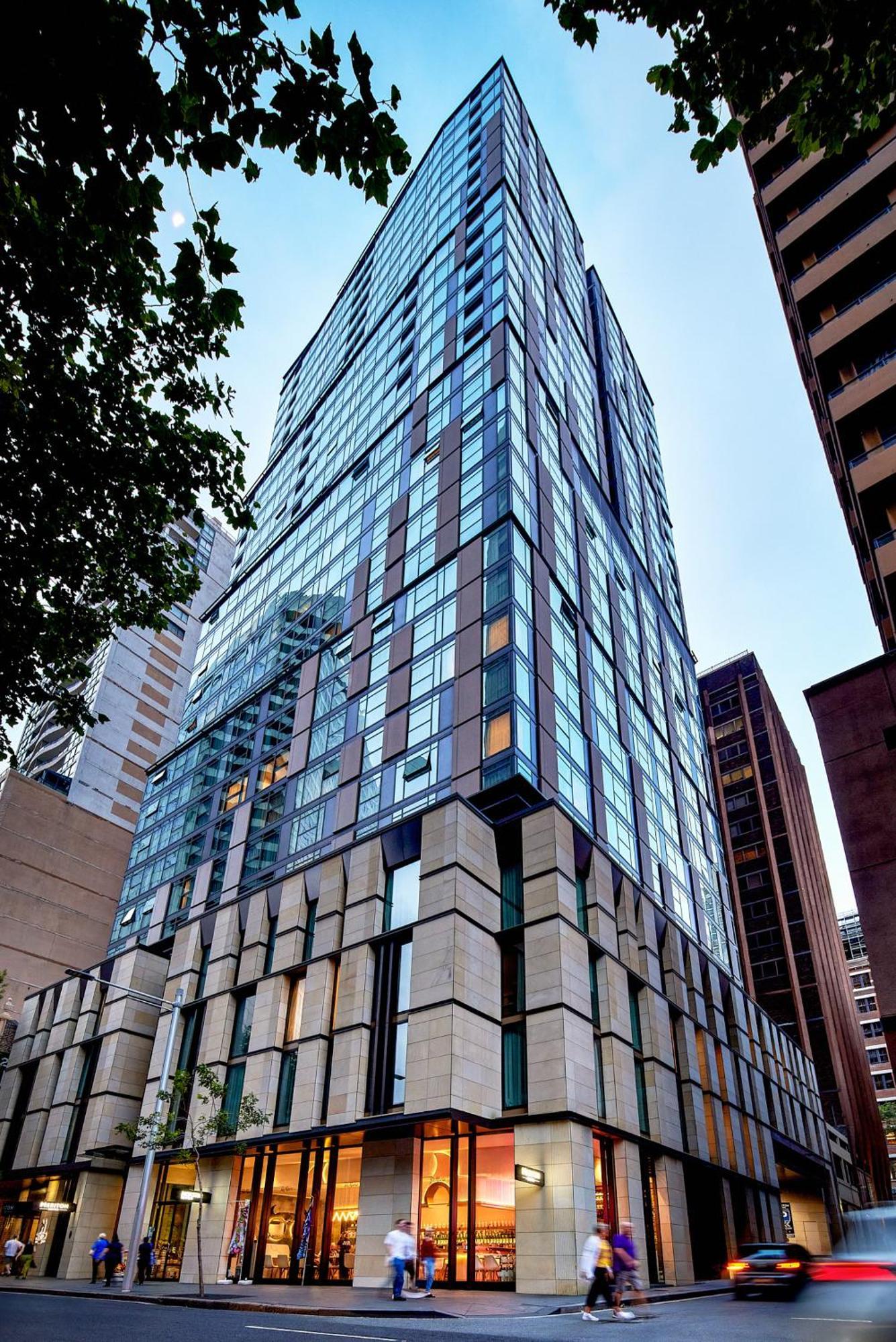 Meriton Suites Sussex Street, Sydney Exterior photo The photo shows a modern high-rise building with a sleek, glass facade reflecting its surroundings. The structure features a combination of glass and concrete materials, with multiple stories visible. The lower part of the building has a sturdy, text